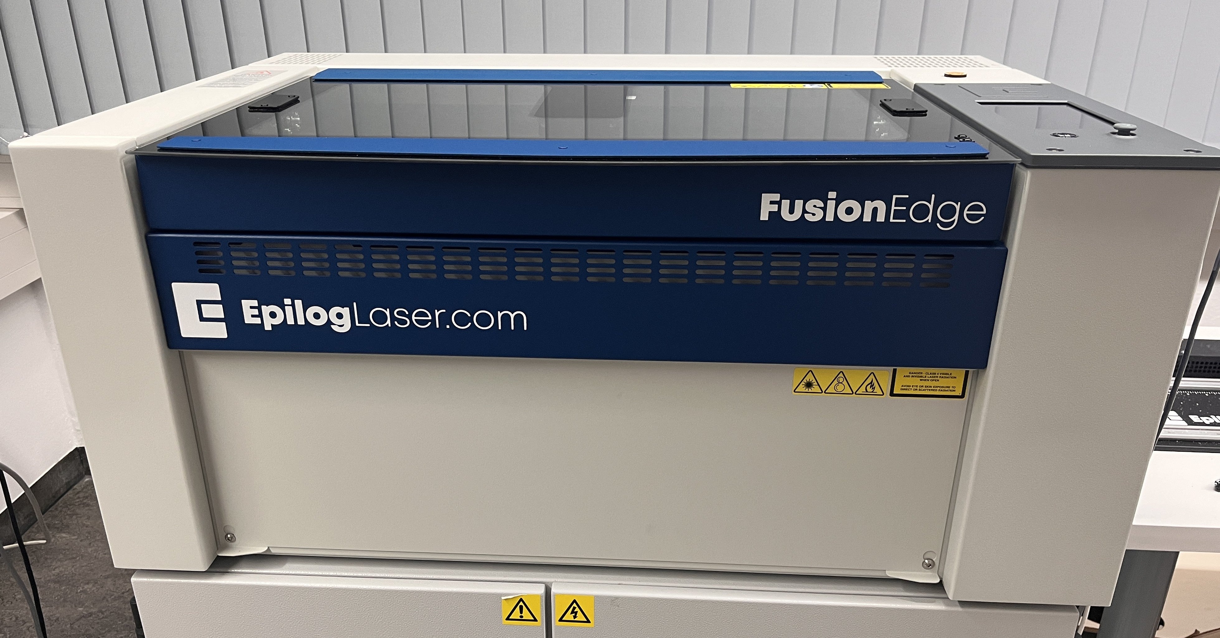 Epilog Fusion Edge 12 Maschine in Blau und Weiß mit Schneidtisch, Glasdeckel und Touchscreen auf einer Absauganlage.