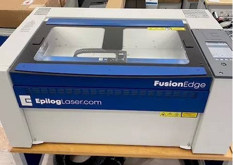 Epilog Fusion Edge 12 Maschine in blau und weiß mit Schneidbrett, Gravierkopf, Touchscreen, Glashaube, Lüftern und Warnhinweisen.
