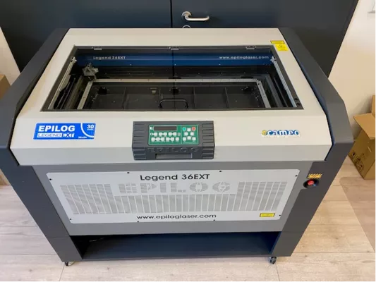 Epilog E9060 Legend 36 EXT Maschine in weiß und schwarz mit Schneidbrett, Glashaube, Einstellungsmenu, Not-Aus Knopf und Rollen am Boden.