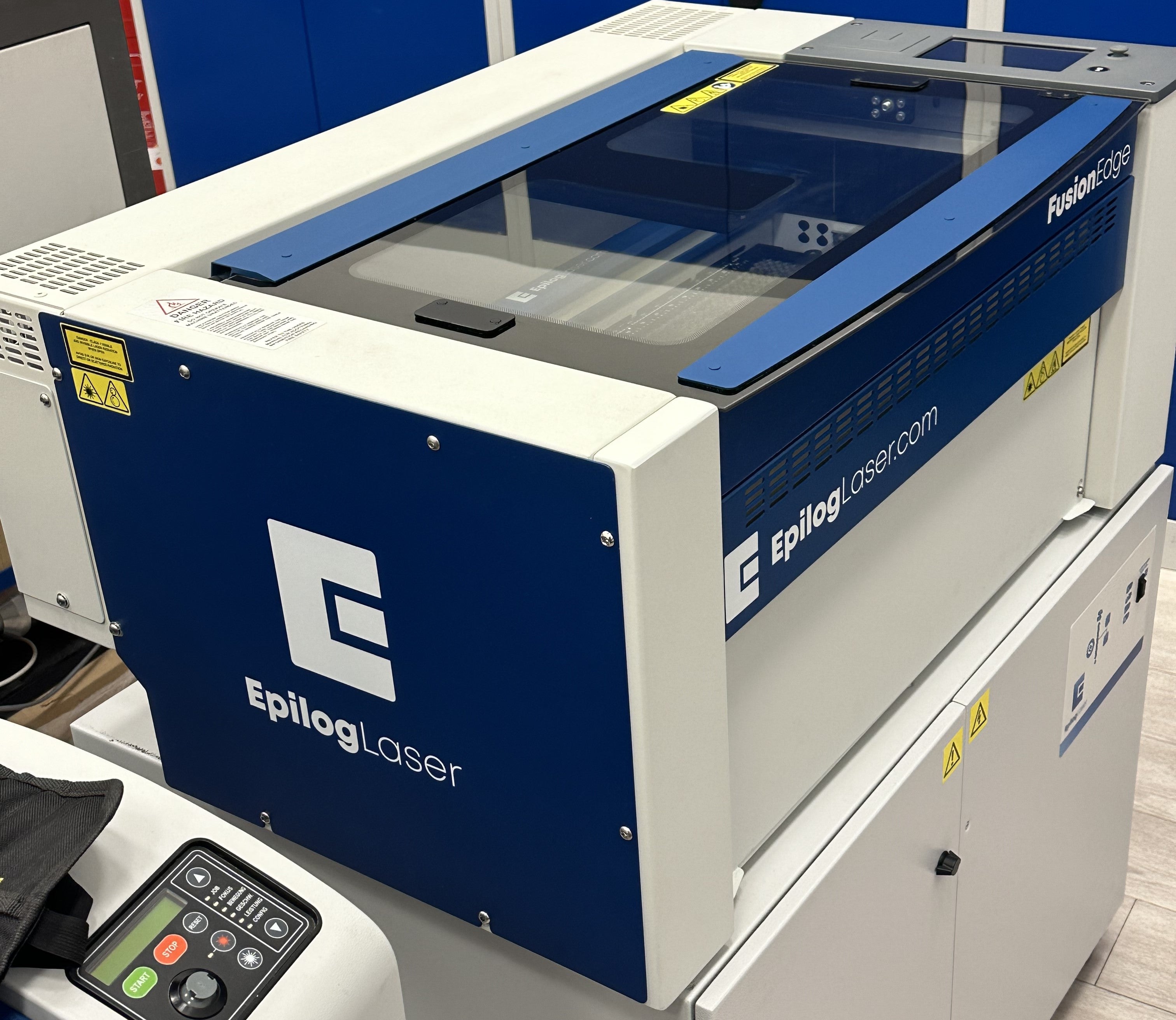 Epilog Fusion Edge 12 Maschine in Blau und Weiß mit Schneidtisch, Glasdeckel, Touchscreen und Lüftern auf einer Absauganlage.