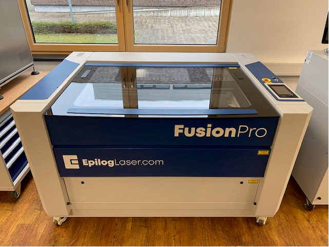 Epilog Fusion Pro 48 Maschine in Blau und Weiß mit Schneidtisch, Glashaube, Lüftern, Touchscreen, Not-Aus Knopf und Rollen am Boden.