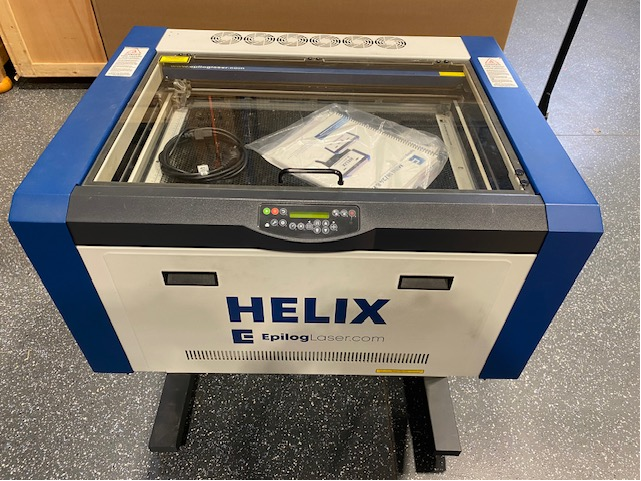 Epilog Helix Maschine in Blau und Weiß mit Schneidtisch, Netzkabel, Glashaube, Lüftern, Einstellungsmenu und Rollen am Boden.