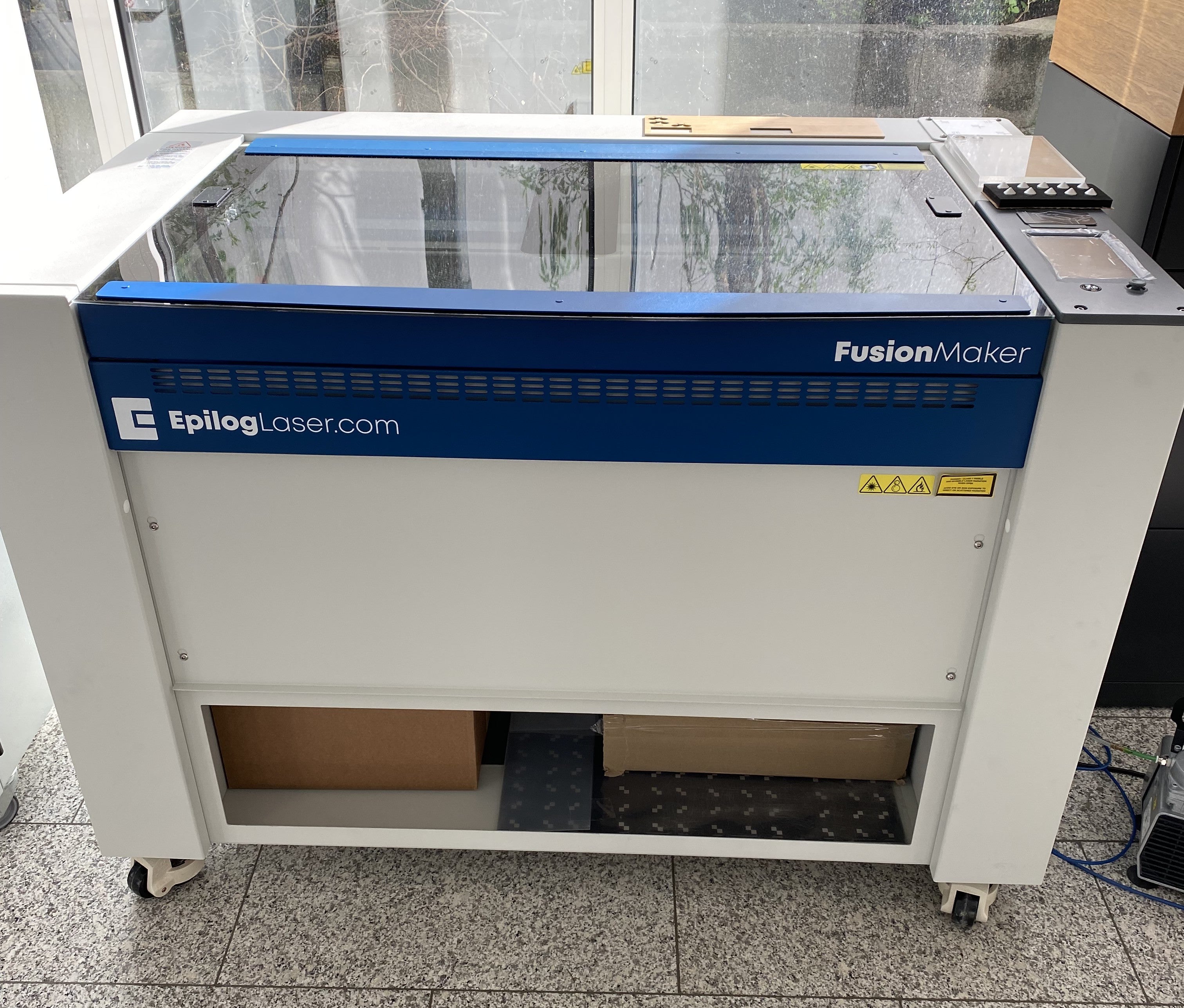 Epilog Fusion Maker 36 Maschine in Blau und Weiß mit Schneidtisch, Glasdeckel, Touchscreen und Rollen am Boden.