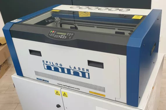 Epilog Mini 24 Maschine in weiß und blau mit Schneidbrett, Glashaube, Lüftungen, Einstellungsmenu und Warnhinweisen.